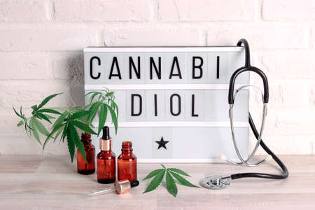Bottles with CBD oil, droppers, lightbox, stethoscope and marijuana leaves on a white brick wall....