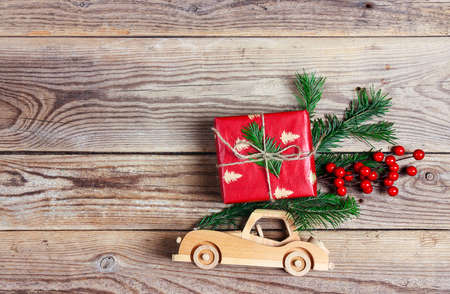 Wooden toy car with gift box and fir branch on the roof on wooden background. Top down Christmas composition with copy space.の写真素材