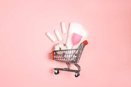 Shopping cart with menstruation period accessories on pink background. Sanitary pads, tampons, menstrual cup and cotton bolls. Flat lay, top view with copy space.の写真素材