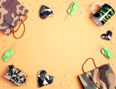 Frame of camouflage hearts, gifts, bag, mug and origami paper tanks on yellow background. Top down...
