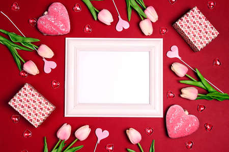 Empty white photo frame with tulips flowers, gifts and hearts on red background. Copy space, flat...