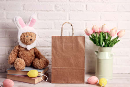 Brown paper shopping bag, funny toy bear with rabbit ears, tulips and colored eggs on a table...
