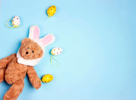 Easter background with a funny toy bear with rabbit ears and colored eggs on blue background. Copy space for text.の写真素材