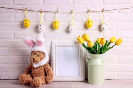 Empty photo frame with a funny toy bear with rabbit ears, tulips and colored eggs against a white...