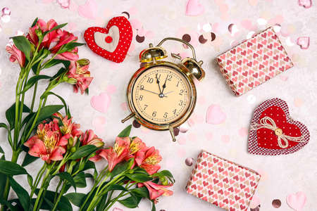 Golden alarm clock with gifts, alstroemeria flowers, hearts and confetti on light marble table....