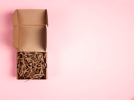 Open brown box with brown shredded paper on pink background. Paper gift box with decorative straws fillers for your product placement. Copy space for produce and text.の写真素材