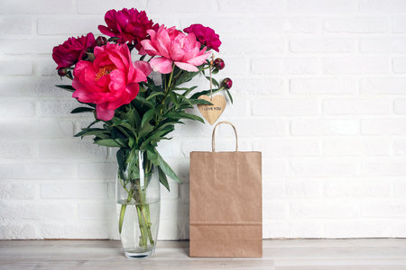 Purple peony flowers bouquet with paper shopping bag on white brick wall. Copy space for text. Shopping and sale concept.の写真素材