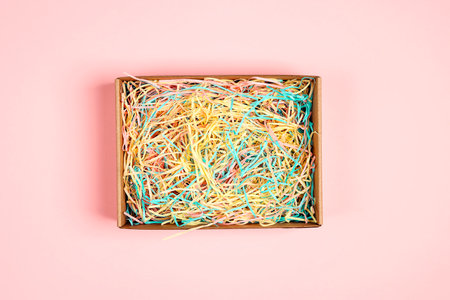 Open brown box with colorful pastel shredded paper on pink background. Paper gift box with decorative straws fillers for your product placement. Flat lay, top view. Center composition with empty space for produce.の写真素材