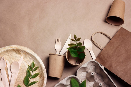 Eco-friendly packaging and tableware set on brown paper background with green leaves and copy space. Bamboo plates, wooden cutlery, paper cups, container and bag. Zero waste, plastic free, biodegradable, disposable tableware. top view.の写真素材