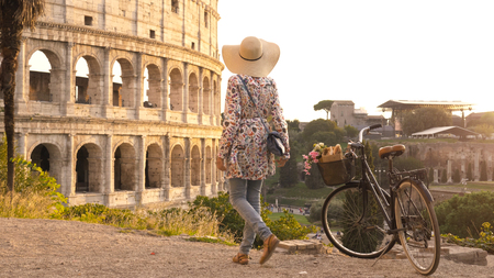 Young woman tourist with bycicle takes pictures of the colosseum in rome at sunset with smartphone. Beautiful stylish dress with large hat, flowers and bread in basket.の写真素材