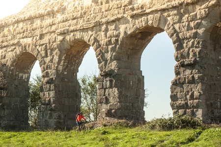 Cyclist riding bike on dirt road under ancient roman aqueduct in a park. Young attractive athletic man with orange sportswear and backpack in parco degli acquedotti in Romeの写真素材