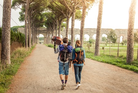 Young couple backpackers tourists with guitar walking towards roman aqueduct ruins in acquedotti park in rome.の写真素材