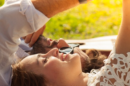 Close-up happy beautiful girl with white dress and attractive man with sunglasses taking selfies with smartphone lying on colorful blanket on grass. Young couple with guitar on sunny summer day.の写真素材