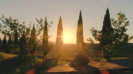 Young woman sitting on old vintage cart looking at sunset through cypress trees in Italian tuscany landscapeの写真素材