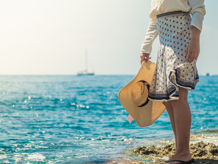 Young woman standing on rocky cliff in Palmarola island in front of the ocean on a sunny day. Elegant white dress with skirt and hat.の写真素材