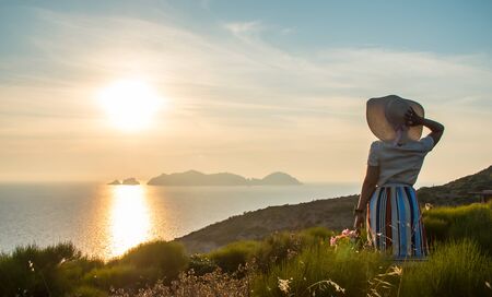 Young woman with flowers basket enjoying sunset view on the hill over the seaside. Large hat, fashion white shirt, colorful skirt.の写真素材
