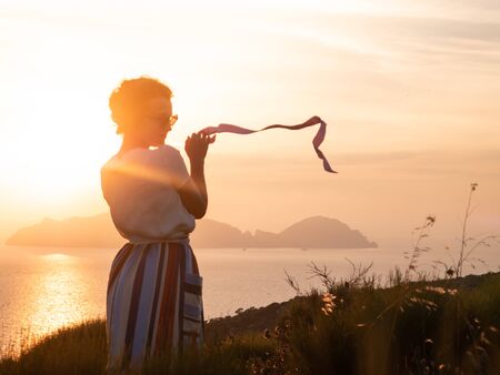 Young woman waving pink ribbon in front of sunset view on the hill over the seaside. Fashion white shirt, colorful skirt, sunglasses.の写真素材