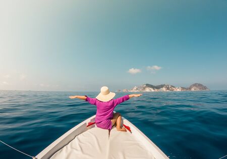 Young woman sitting on bow of boat in the middle of the sea. Happy girl with hat raising hands enjoying freedom travel.の写真素材