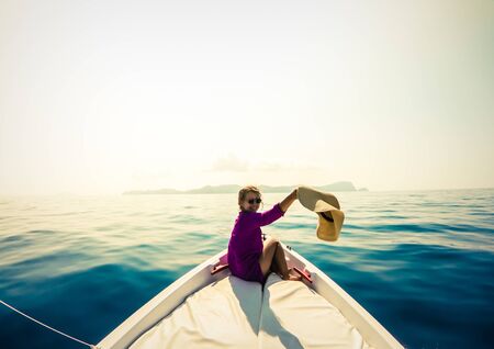 Young woman sitting on bow of boat in the middle of the sea. Happy girl with hat raising hands enjoying freedom travel.の写真素材