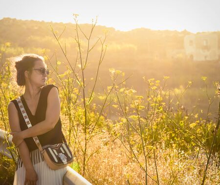 Young woman in green nature at sunset on Ponza Island Italy. Fashion black dress, purse and sunglasses. Summer evening.の写真素材