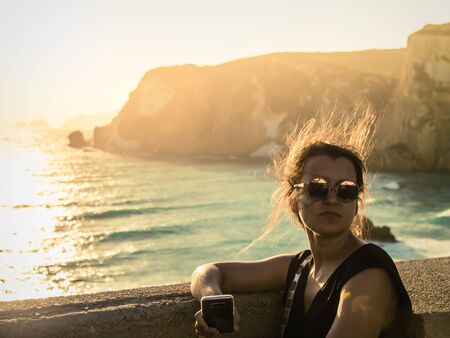 Young woman sitting at sunset in front of the sea on Ponza island coast. Hair in the wind, fashion sunglasses.の写真素材