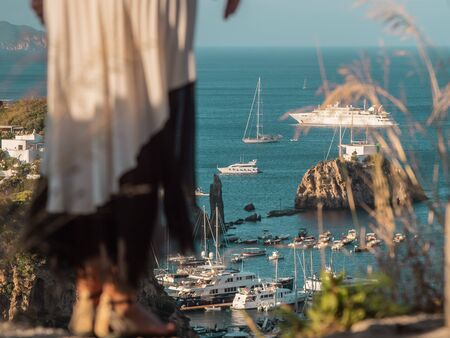 Happy young woman tourist looking from hill at the sea landscape with boats on Ponza island in Italy. Low angle close up. Fashion black and white skirt.の写真素材