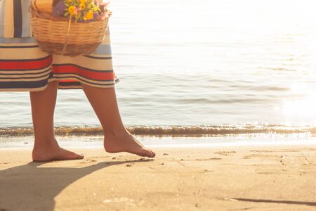 Young woman walking barefoot on the beach in Ponza Island harbor, Italy. Fashion white shirt and colorful skirt, basket with flowers, book and wine bottle.の写真素材
