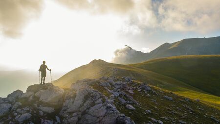 Young hiker man with backpack and walking poles, standing on peak of a mountain looking at sunset in...