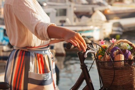 Young woman with bike at Ponza Island harbor pier in Italy. Tourist with large hat, fashion shirt and colorful skirt. Basket with wine and flowers in front of shops and boats.の写真素材