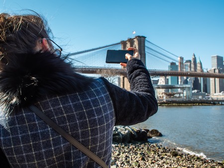 Young Woman tourist by the river in Dumbo taking pictures of Brooklyn Bridge and Cityscape of New York skyline.の写真素材
