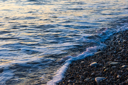 sea pebble beach with multicoloured stones, transparent waves with foam, on a warm summer dayの写真素材