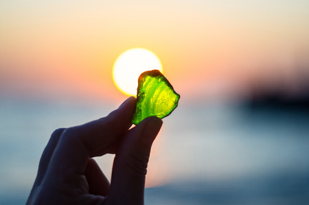 the sea smoothed green glass in hand on background of sunsetの写真素材