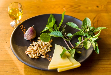 Fresh ingredients for preparing Italian pesto sauce - lemon basil sprigs, peeled seeds of cedar nuts, large garlic clove, Greek olive oil, Parmesan cheese, on a ceramic plate, in scattered sunlightの写真素材