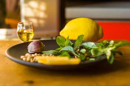 Fresh ingredients for preparing Italian pesto sauce - lemon basil sprigs, peeled seeds of cedar nuts, large garlic clove, Greek olive oil, Parmesan cheese, on a ceramic plate, in scattered sunlightの写真素材