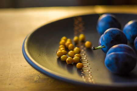 still life - juicy blue plums and sea buckthorn berries in a ceramic plate on a wooden background, close-upの写真素材