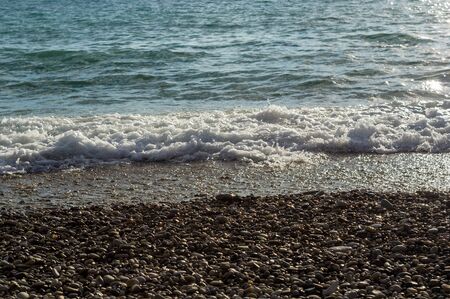pebble stones on the sea beach on a warm summer day, the rolling waves of the blue sea with white foamの写真素材