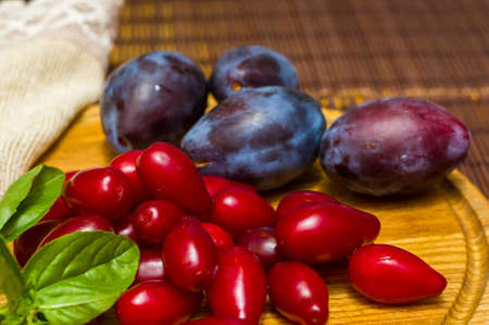 Still life - fresh large dogwood berries, purple plums and a sprig of basil on a wooden board, close upの写真素材