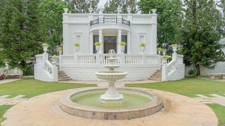 Location photos A three-tiered, circular white fountain with a stone tile floor. In the middle of the green grass The back is a luxurious European-style building decorated with vasの写真素材