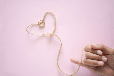 Heart shaped rope and gold ring with blur image of woman finger on pink paper background. romantic and love valentines day concept. flat lay with copy space.の写真素材