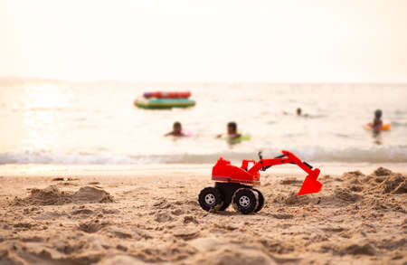 Toy tractor on sand beach at coast with child playing on sea blured. sun set on ocean. tourist in...