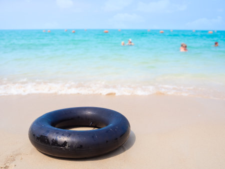 Black swim ring or inflatable rubber toy on sand beach at coast with blured people swimming in blue sea. accessories for tourist vacation summer travel in holidays.の写真素材