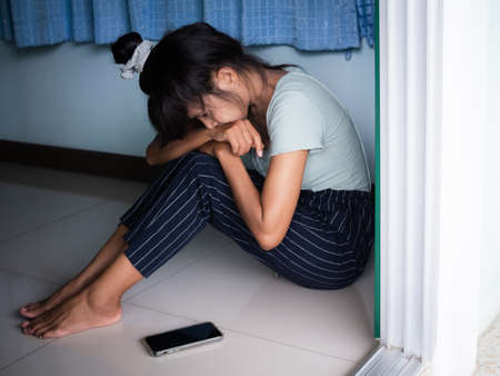 Broken Heart concept. Alone woman sitting and looking mobile fone at room. She sad, cry, stress and lonely. card or poster for problem couple disappointed in love, pain.waiting for call from husband.の写真素材