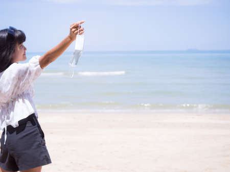 Woman holding medical mask at coast on blur blue sea and blue sky background.lifestyle happiness vacation freedom. tourist tropical travel summer holidays nature concept. authentic skin tan,slim fit.の写真素材