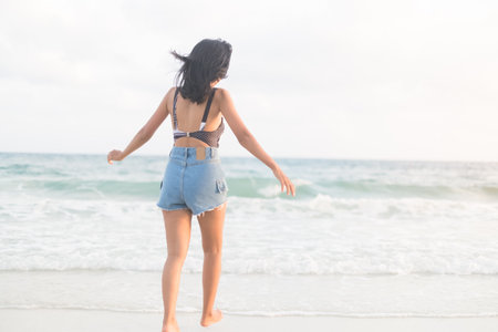Portrait Happy Girl Running on Sand Beach with Blue Sea,Slim Fit Women with Short Jeans Stand at Ocean Coast,Authentic Female Skin Tan and Black Hair Asian,Tourism Travel Tropical Summer Holiday.の写真素材