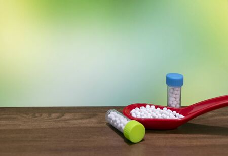 Close view of White pills in red spoon with Homeopathic medicine bottles on wood surface and green mix yellow backgroundの写真素材
