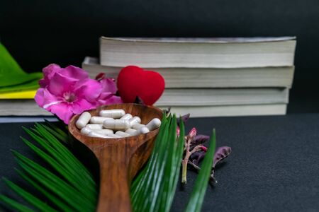 Herbal medicine capsules in wooden spoon with red heart, pink flower, green leaves and books on dark background- Herbal Medicine Conceptの写真素材