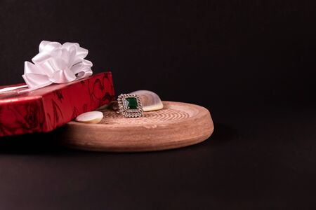 Emerald ring with seashells and decorated red gift box on sand Stone with black background