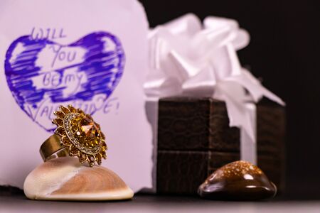 Valentine gift concept. Hand written Valentine quote on white paper with decorated brown gift box and cocktail ring on seashell with black backgroundの写真素材