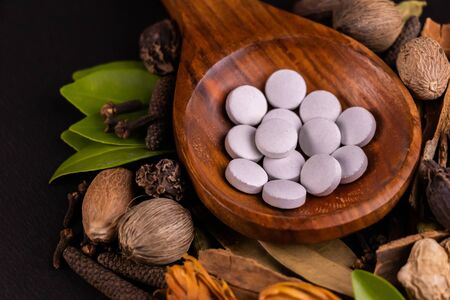High angle closeup shot of white herbal pills in a wooden serving spoon with green and dry herbs on black background, herbal medicine conceptの写真素材