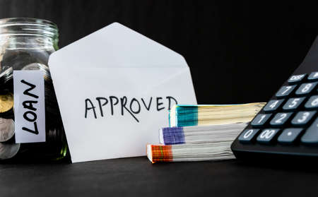 A white opened envelope with the word Approved and calculator, money, coins, glass jar aside
